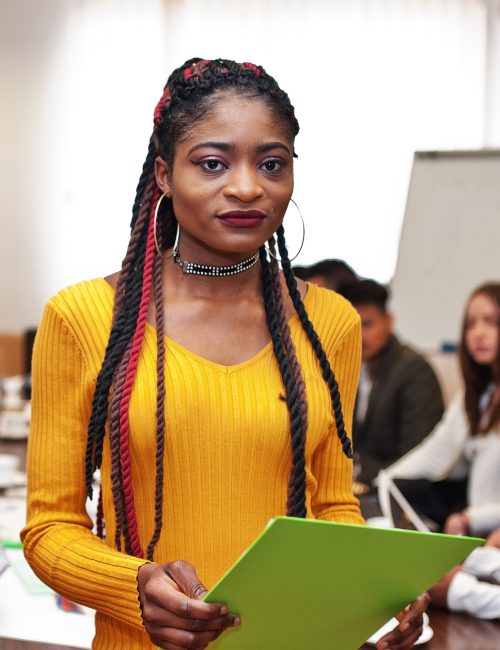 Face of handsome african business woman in yellow dress, holding clipboard on the background of business peoples multiracial team meeting, sitting in office table.